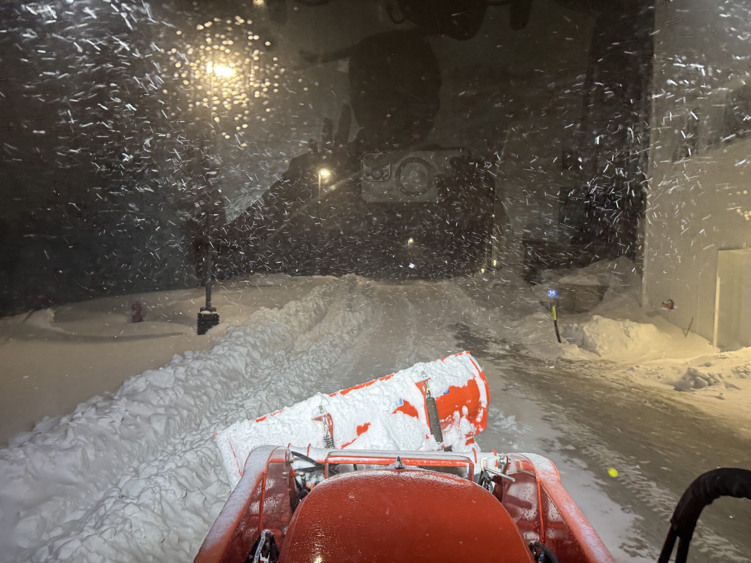 Looking out the window of a tractor with a snow plow blade, at night. It is snowing and there are 3 foots snowbanks.