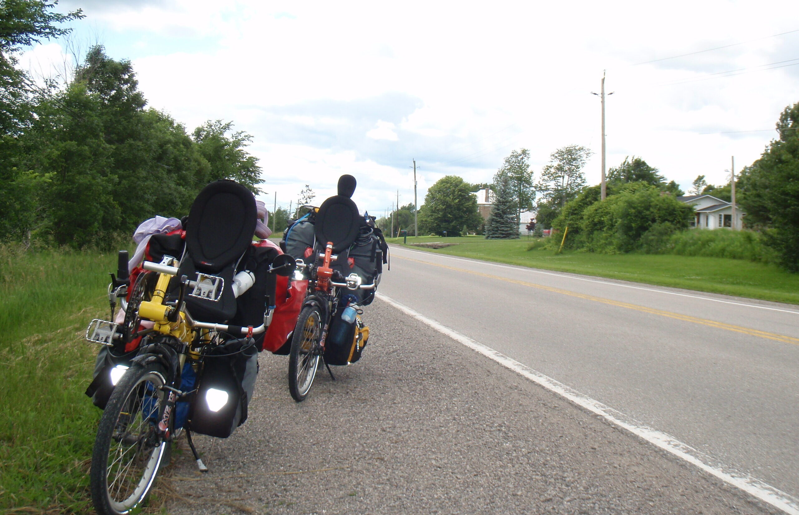 Two loaded recumbent touring bicycles are parked on the gravel shoulder of a quiet rural road. The bikes are equipped with panniers, bags, and gear for long-distance travel. Trees and grassy fields line both sides of the road, with a few houses visible in the distance under a bright, cloudy sky.