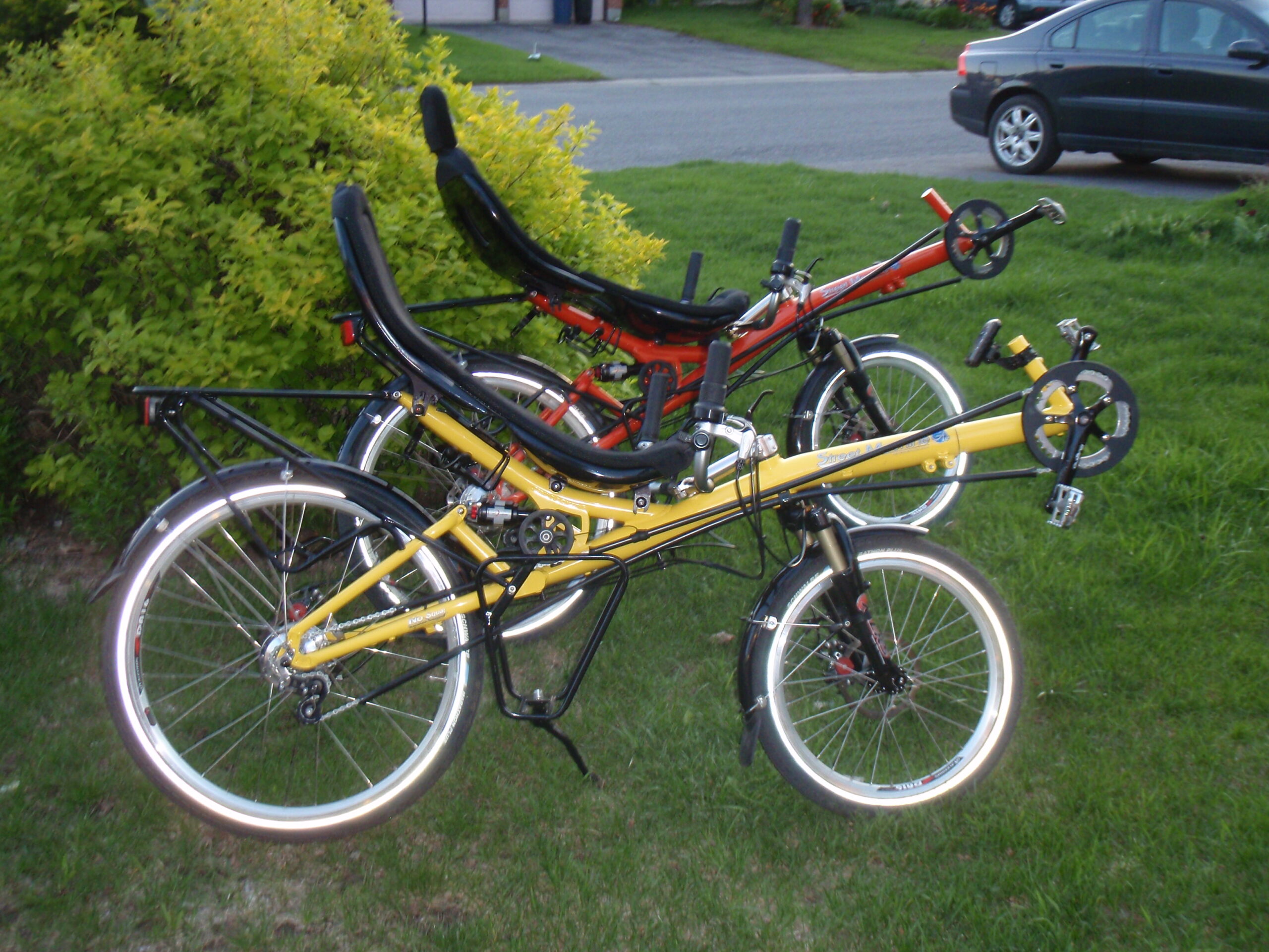 Two recumbent bicycles are parked on a grassy lawn beside a hedge. A yellow recumbent bike with a short wheelbase stands in the foreground, while an orange recumbent bike is positioned behind it. Both bicycles have low-slung seats with backrests and are angled to the right. A residential street, driveway, and parked car are visible in the background.