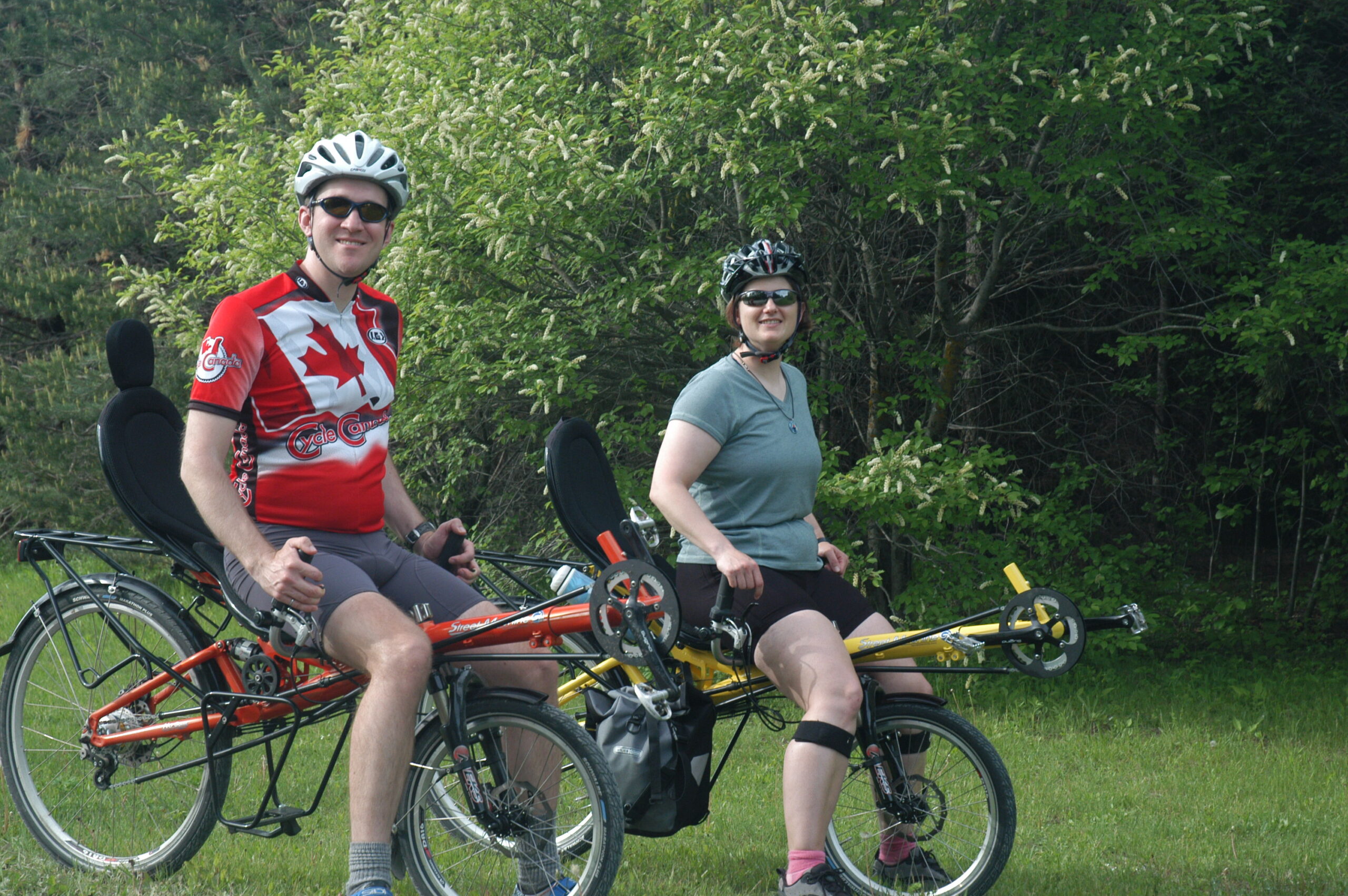 Two people wearing bicycle helmets and sunglasses sit on recumbent bicycles on a grassy area beside a wooded roadside. One bicycle is orange and the other is yellow, both HP Velotechnik Streetmachine models with upright seats and touring racks. The male rider on the orange bicycle wears a red cycling jersey with a large maple leaf design, while the female rider on the yellow bicycle wears a gray shirt and black cycling shorts. Dense green trees and shrubs fill the background.
