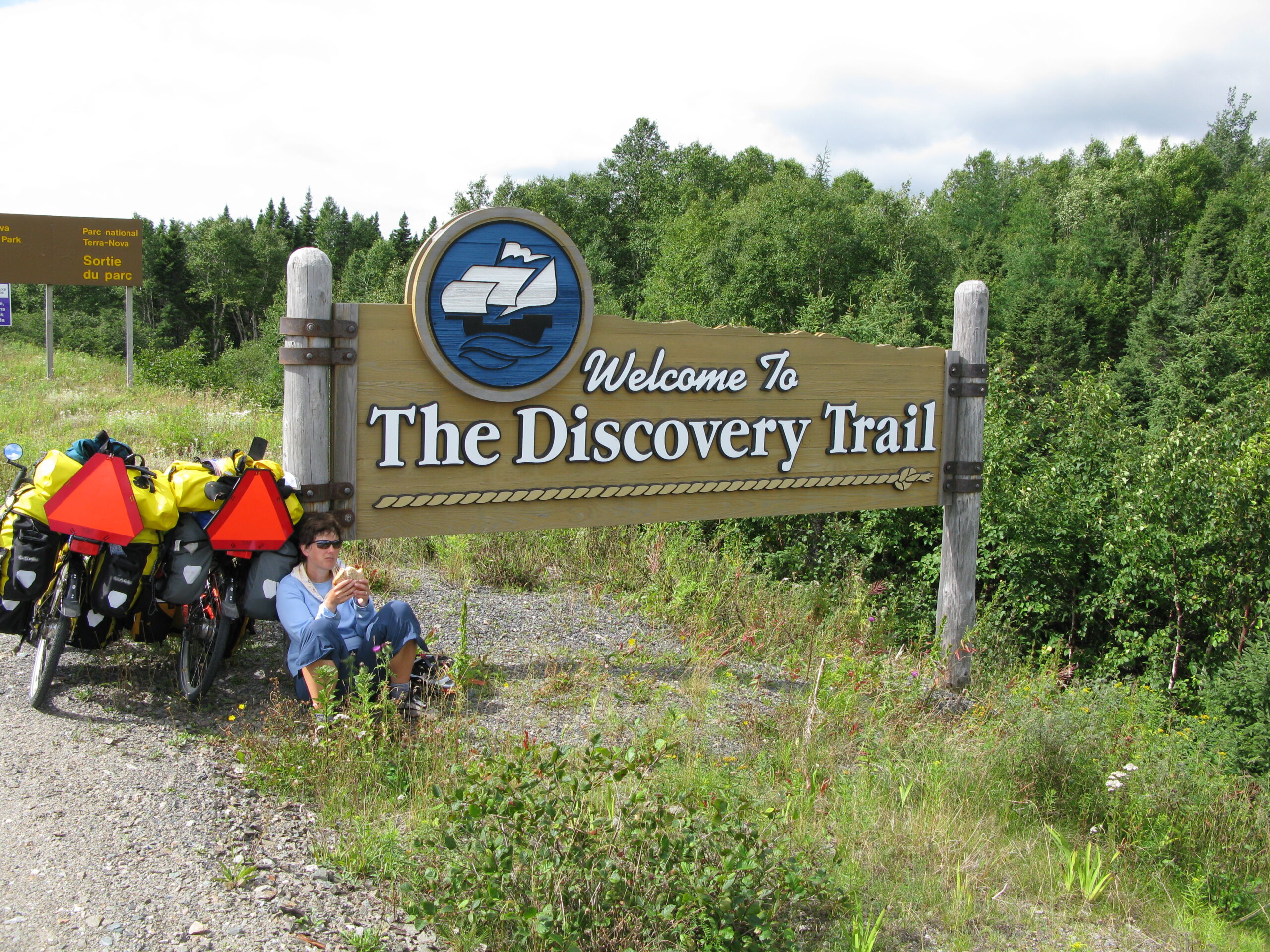 A touring cyclist sits beside a heavily loaded bicycle beneath a wooden “Welcome to The Discovery Trail” sign. A nearby Parks Canada sign marks the exit from Terra Nova National Park, with forest behind.